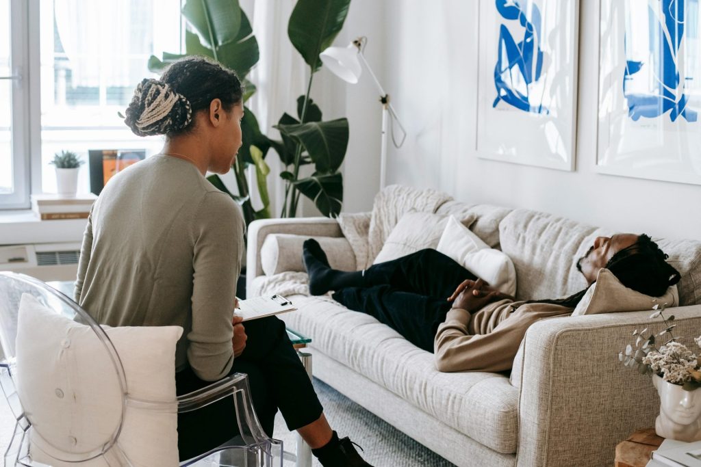 Ethnic female therapist with clipboard sitting near male client lying on couch with clasped hands during psychotherapy session in cozy office
